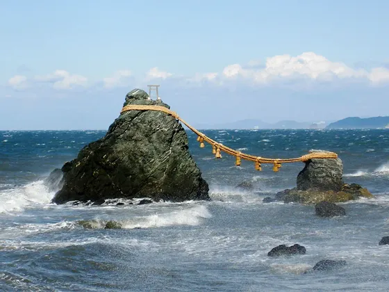 Due grandi faraglioni in un oceano agitato sono collegati da una spessa corda. La roccia più grande è sormontata da un piccolo torii.