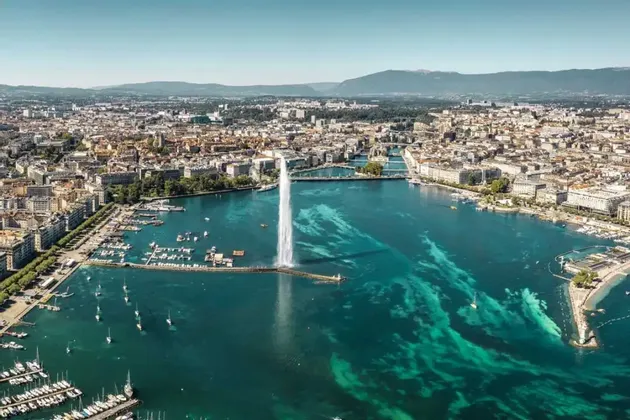 Una veduta aerea di una grande fontana d'acqua che zampilla da un lago turchese, con un paesaggio urbano e un porto pieno di barche lungo la riva.