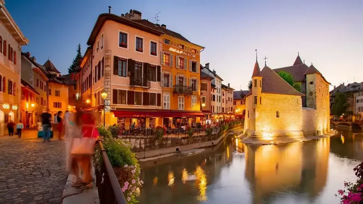 Un histórico edificio de piedra se asienta en un canal, rodeado de restaurantes y tiendas iluminadas a lo largo de una calle adoquinada al anochecer.