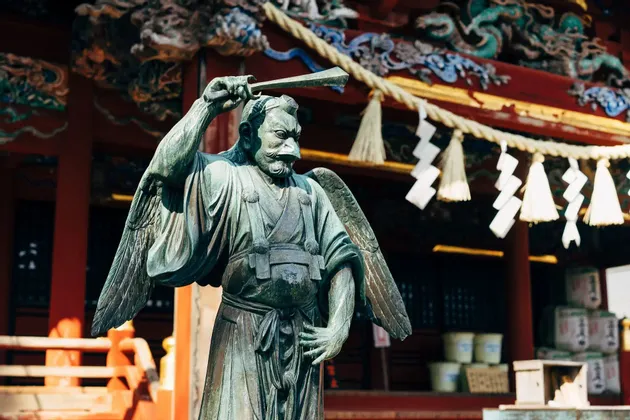 Una statua in bronzo di un Tengu alato, divinità giapponese con un becco prominente, si erge all'esterno di un tradizionale tempio rosso.
