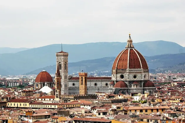 La cupola e il campanile di una grande cattedrale si ergono sopra i tetti rossi di una città, con le montagne blu visibili in lontananza.
