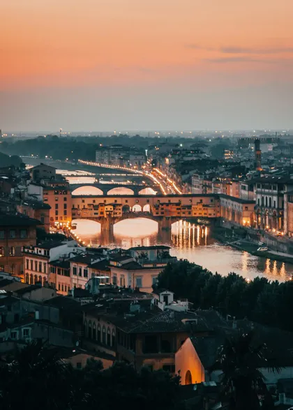 Un ponte illuminato con molteplici archi attraversa un fiume in uno scenario cittadino al tramonto, con le luci che si riflettono sull'acqua.