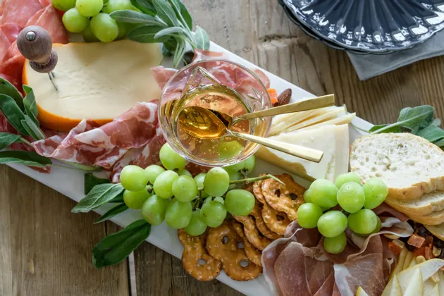 Una vista dall'alto di un tagliere di salumi e formaggi con formaggi assortiti, salumi, uva verde, pretzel e pane a fette su un tavolo di legno.