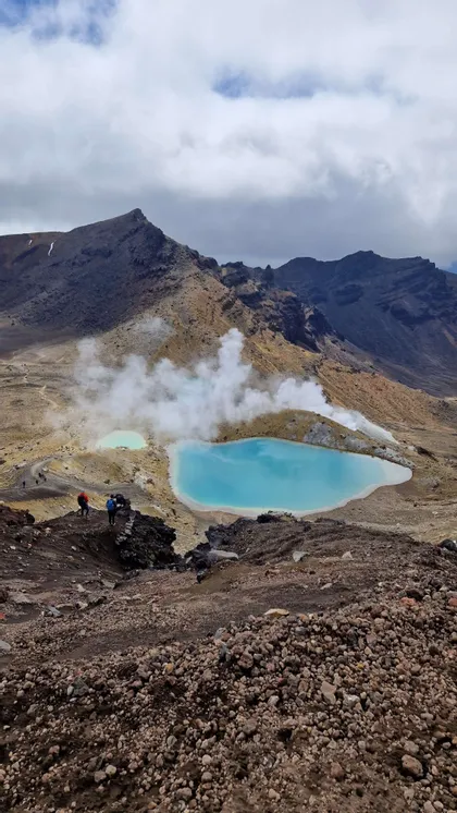 Eine WeRoad Gruppenreise mit Wanderern auf einem felsigen Pfad, der zwei leuchtend blaue Geothermal-Seen überblickt, aus denen in einem Vulkantal Dampf aufsteigt.