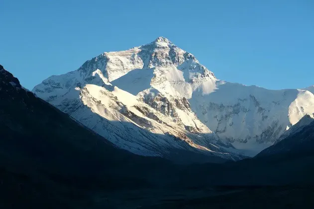 Una cima montuosa alta e innevata è illuminata dal sole contro un cielo blu limpido, sorgendo da una valle in ombra.