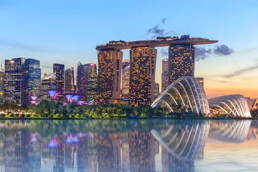 A modern city skyline with illuminated skyscrapers and glass domes reflecting in the water at dusk.
