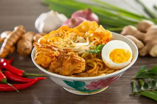A bowl of curry noodle soup with chicken, tofu, and a hard-boiled egg, surrounded by fresh ingredients like ginger and red chilies.