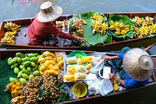 An overhead view of two vendors in conical hats selling fresh tropical fruits like bananas and mangoes from their boats at a floating market.