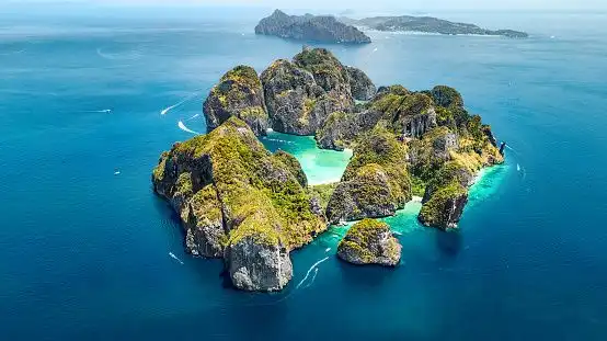 An aerial view of rocky, green-covered islands in a turquoise sea, with several small boats creating wakes in the water.