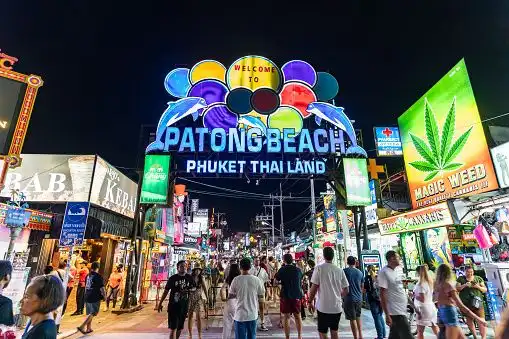 A large, colorful neon sign for a beach destination arching over a crowded street filled with people at night.