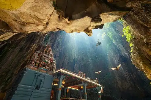 An ornate temple built inside a large cave, with sunbeams shining down from an opening as birds fly past.