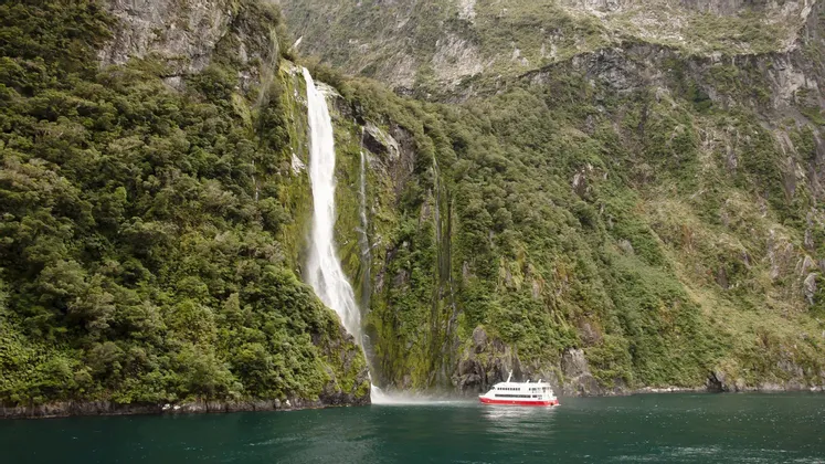 Ein rot-weißes Ausflugsboot fährt auf dem Wasser am Fuße eines hohen Wasserfalls an einer üppigen, felsigen Klippe.