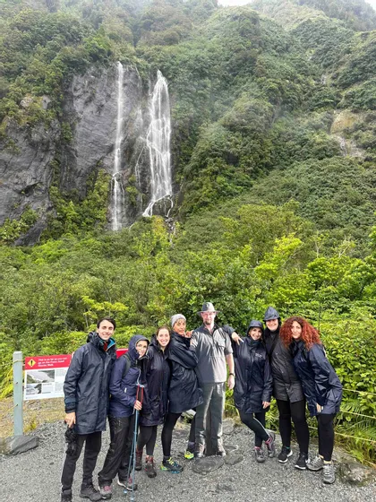 Eine WeRoad Gruppenreise posiert in Regenmänteln für ein Foto vor einem hohen Wasserfall in einem üppig grünen Tal.