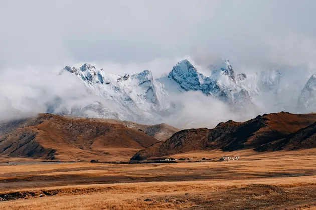 Una vasta pianura dorata con un piccolo insediamento di yurte di fronte a grandi montagne innevate parzialmente coperte da nuvole.