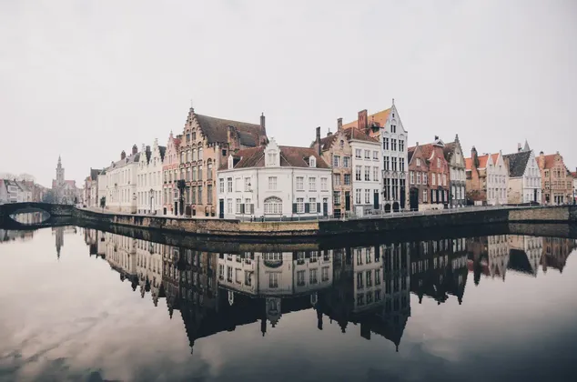 Una hilera de edificios históricos con tejados a dos aguas se alza a lo largo de un canal, con sus fachadas perfectamente reflejadas en el agua tranquila bajo un cielo nublado.