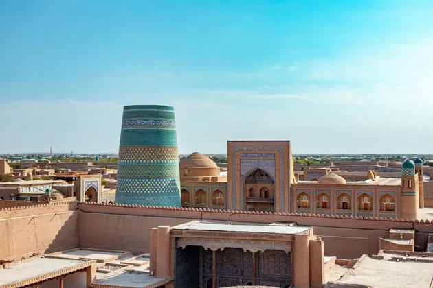 Uno scenario urbano con un grande e ornato minareto dalle piastrelle turchesi che si innalza sopra edifici e cupole color sabbia, sotto un cielo azzurro e limpido.