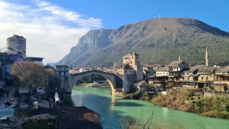 Eine Steinbogenbrücke überspannt einen türkisfarbenen Fluss, der durch eine Stadt mit Steingebäuden und Türmen fließt, mit einem großen Berg im Hintergrund.