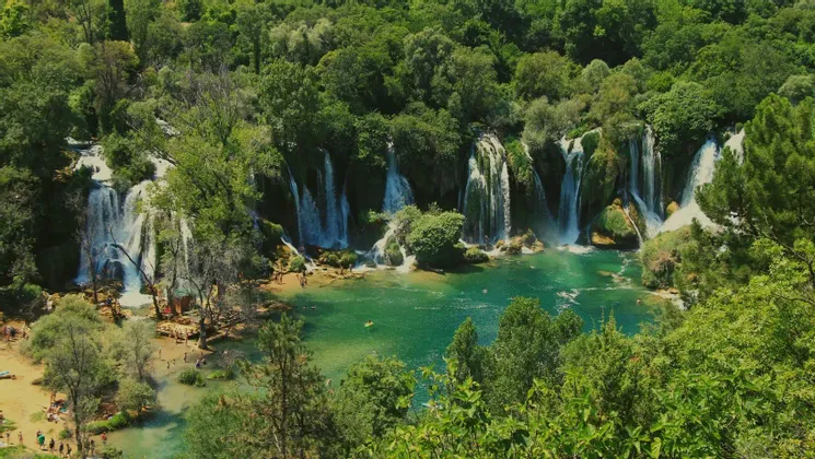 Breite Wasserfälle stürzen in einen türkisfarbenen Pool, in dem Menschen schwimmen, umgeben von dichtem grünem Wald.