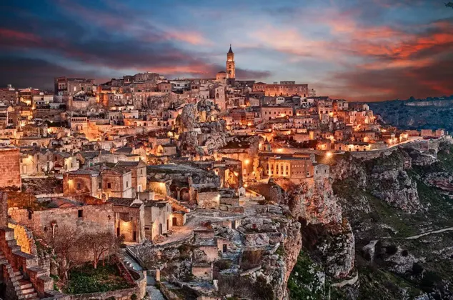 Un'antica città sulla scogliera con edifici in pietra illuminati da luci calde sotto un cielo colorato al tramonto.
