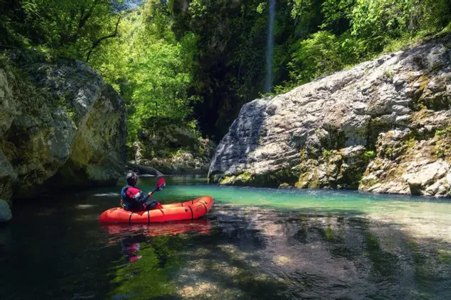 Una persona su un kayak rosso gonfiabile pagaia su un fiume limpido attraverso una gola rigogliosa e rocciosa con una cascata sullo sfondo.