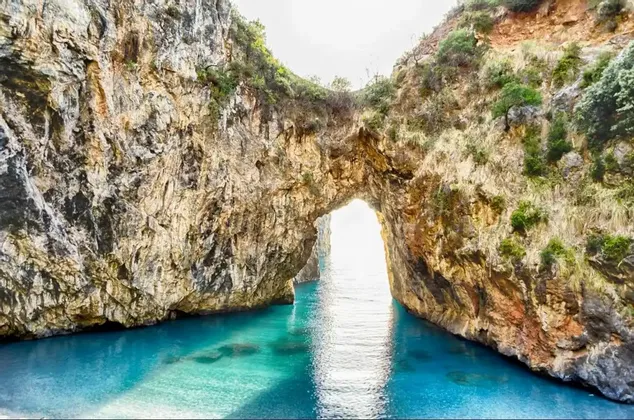 Un grande arco naturale di roccia si erge su acque limpide e turchesi, con il sole che splende luminoso attraverso l'apertura.