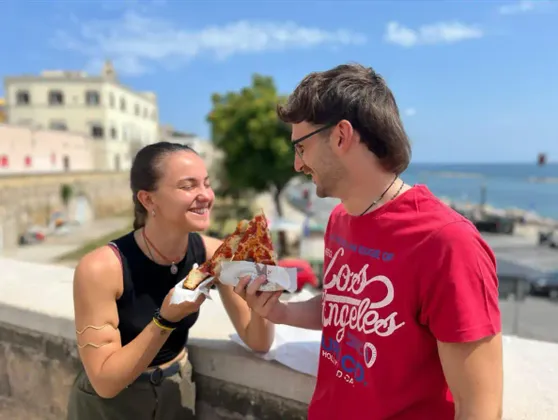 Un uomo con una camicia rossa offre una fetta di pizza a una donna sorridente su una terrazza all'aperto con il mare sullo sfondo.