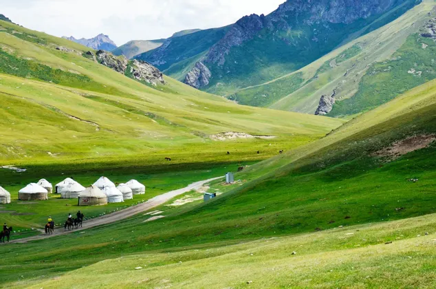 Un viaggio di gruppo WeRoad a cavallo su un sentiero sterrato accanto a un campo di yurte, immerso in una vasta e verde valle di montagna.