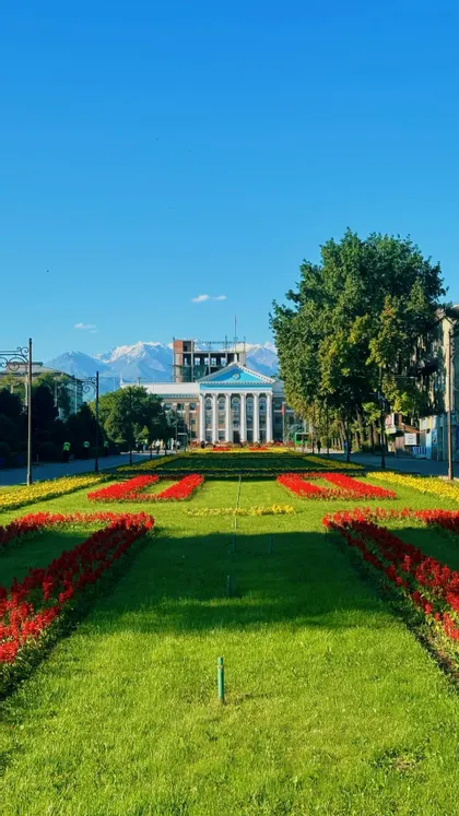 Un parco curato con aiuole di fiori rossi e gialli che conduce a un edificio classico con montagne innevate sullo sfondo.