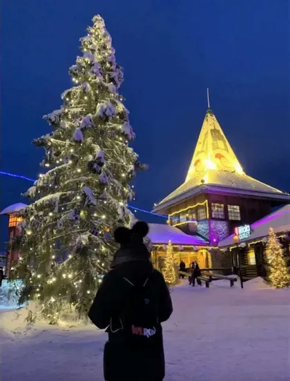 Une personne portant un sac à dos WeRoad regarde un grand sapin de Noël enneigé et illuminé de lumières sur une place de village la nuit.