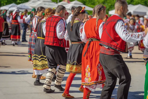 Una fila de bailarines folclóricos con coloridos trajes tradicionales bordados actúan al aire libre.