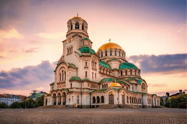 Una gran catedral blanca con cúpulas verdes y doradas se alza en una plaza adoquinada bajo un cielo de atardecer colorido.