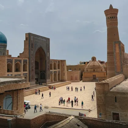 Una vista aerea di persone che camminano in un ampio cortile circondato da storici edifici in mattoni con archi a mosaico blu e un alto minareto.