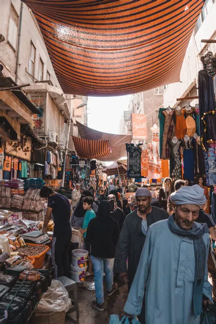 Une foule de personnes traverse un marché de rue animé, bordé d'étals et protégé par un auvent rayé orange.