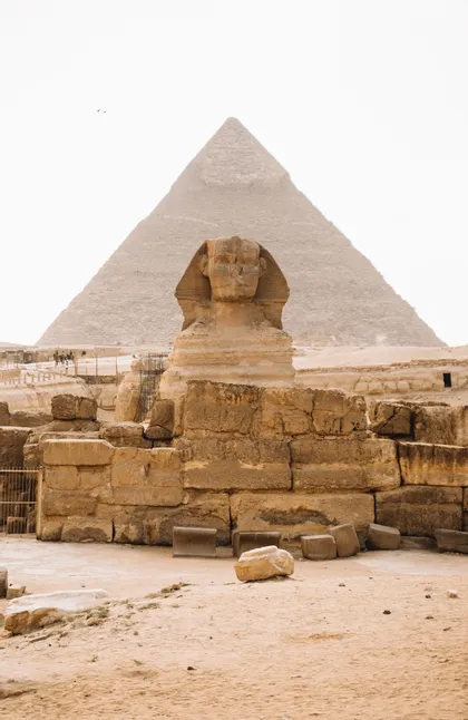 Le Grand Sphinx se dresse devant une grande pyramide dans un paysage sablonneux, avec des ruines de pierre antiques au premier plan.