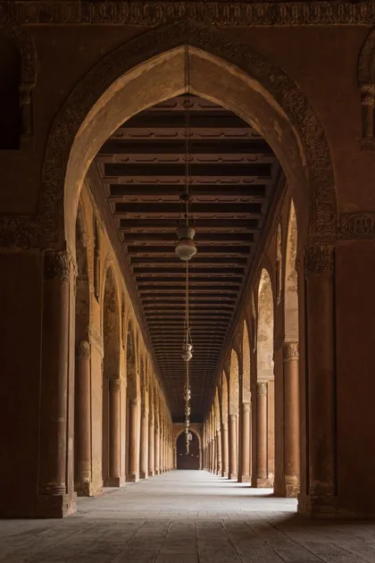 Un long couloir symétrique avec des arches et des colonnes de pierre répétitives, où des lanternes pendent du plafond en bois.