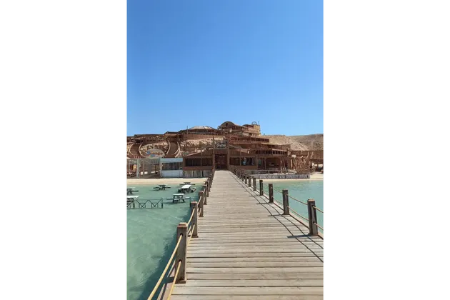Un lungo molo di legno si estende su acque turchesi e limpide verso un grande resort balneare in legno costruito su un versante sabbioso sotto un cielo azzurro e sereno.