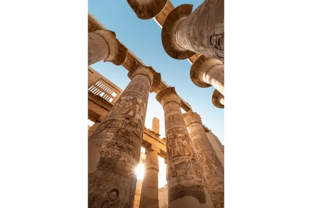 Vista dal basso di imponenti colonne di pietra con geroglifici egizi, con il sole che splende in un cielo azzurro e limpido.