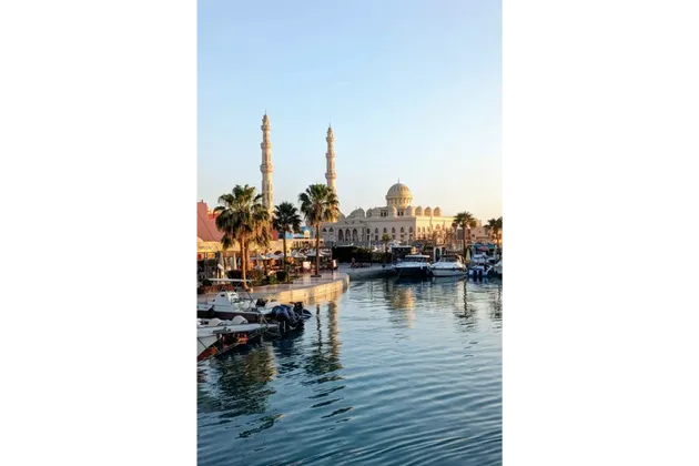 Una grande moschea con due minareti si affaccia su un porto turistico, pieno di barche ormeggiate e palme, sotto un cielo sereno.