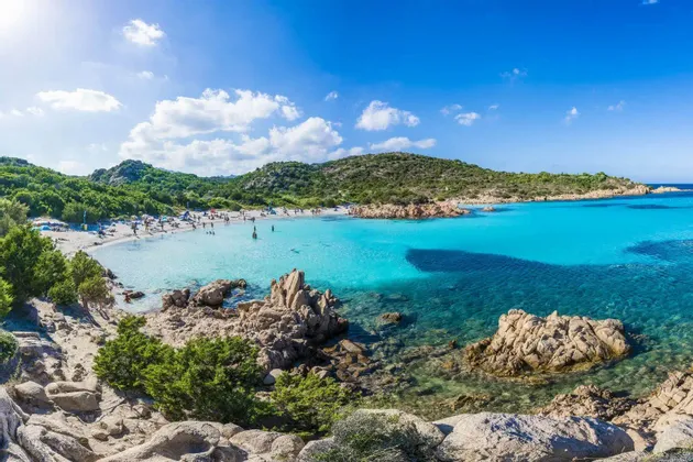 Persone che si rilassano e nuotano in una caletta isolata con acqua turchese, circondata da colline verdi sotto un cielo soleggiato.