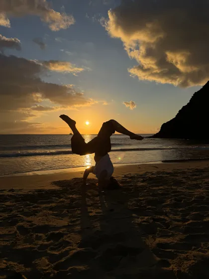 Una persona in silhouette al tramonto, intenta a fare una verticale su una spiaggia sabbiosa con le onde sullo sfondo.