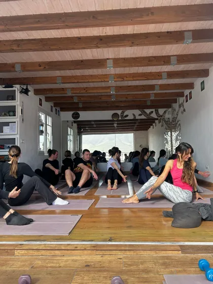 Un gruppo WeRoad in viaggio che partecipa a una lezione di fitness, seduto su tappetini su un pavimento in legno all'interno di uno studio con travi a vista.