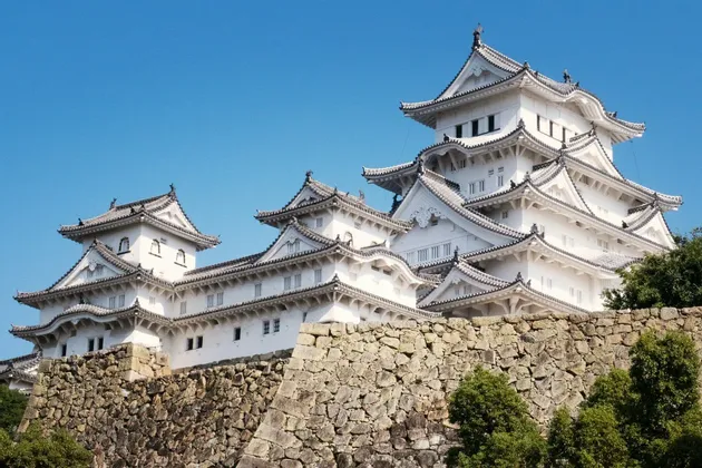 Un tradizionale castello giapponese bianco con tetti a più livelli si erge su un grande muro di pietra sotto un cielo azzurro e limpido.