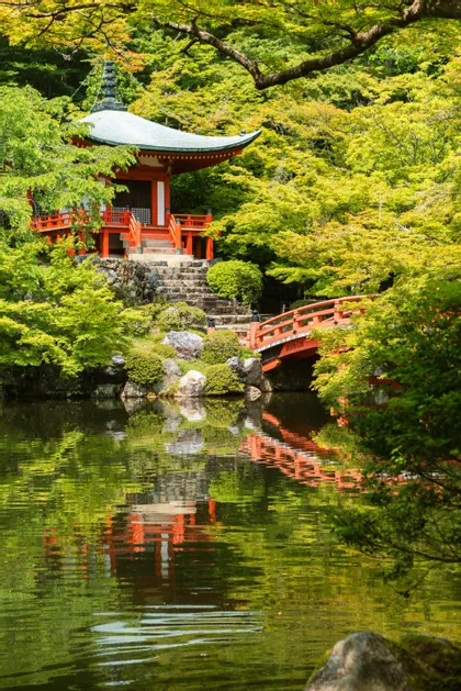 Una pagoda giapponese rossa e un ponticello sono immersi tra alberi verdi lussureggianti, con i loro riflessi che si specchiano in un tranquillo laghetto sottostante.