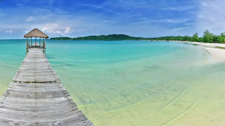 Un ponton en bois avec une cabane au toit de chaume s'avance sur une eau calme et turquoise, à côté d'une plage de sable blanc sous un ciel bleu clair.