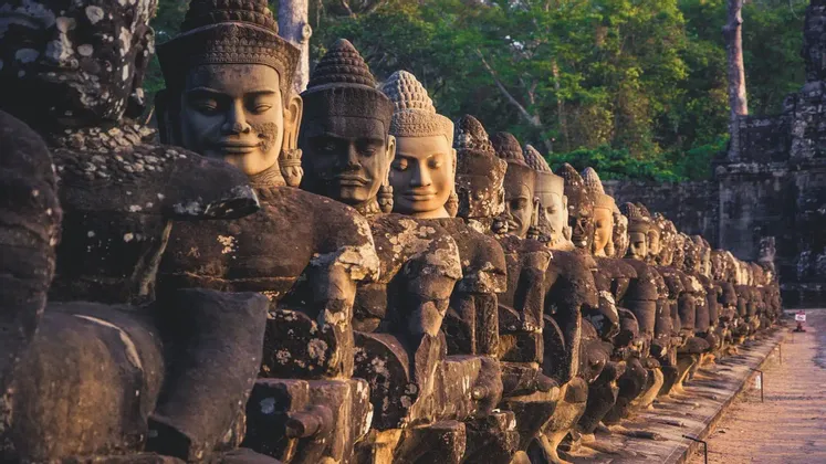 Une rangée de statues de pierre anciennes et érodées, aux expressions sereines, bordant un sentier devant une forêt verdoyante et dense.