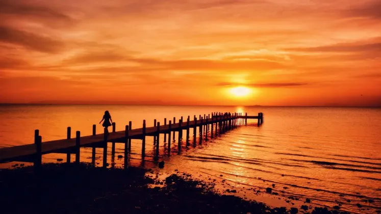 Silhouette d'une personne sur une longue jetée en bois, au-dessus de l'eau, au coucher du soleil orange flamboyant.