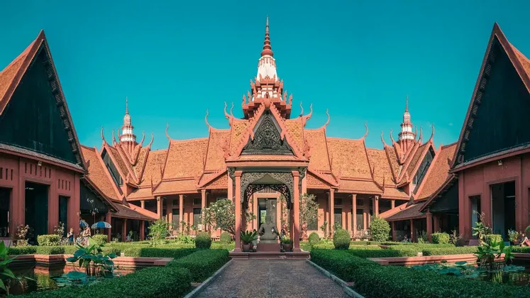 Un bâtiment orné, brun-rougeâtre, à l'architecture traditionnelle entoure une cour centrale avec des jardins manucurés et un chemin de pierre sous un ciel clair.