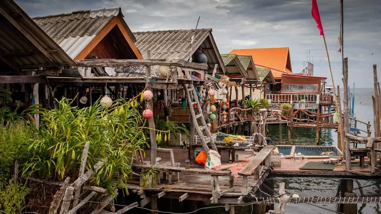 Un ensemble de maisons rustiques en bois sur pilotis construites sur l'eau, décorées de bouées colorées et de plantes sous un ciel nuageux.