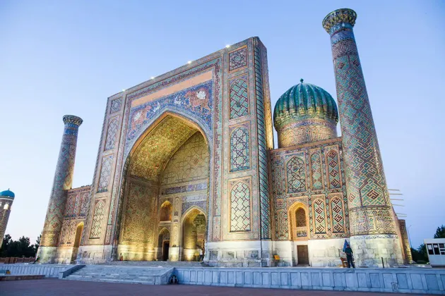 Una madrasa sontuosa con complesse piastrelle blu e oro, un grande arco, una cupola turchese e minareti, illuminata al crepuscolo.