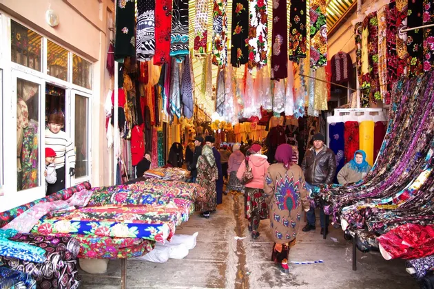 Un vivace corridoio di mercato all'aperto, affollato di persone che curiosano tra bancarelle piene di tessuti e stoffe colorate.